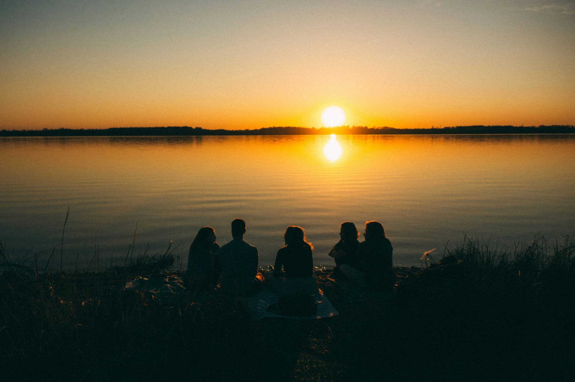 Five people sitting together on a lakeshore watching a golden sunset